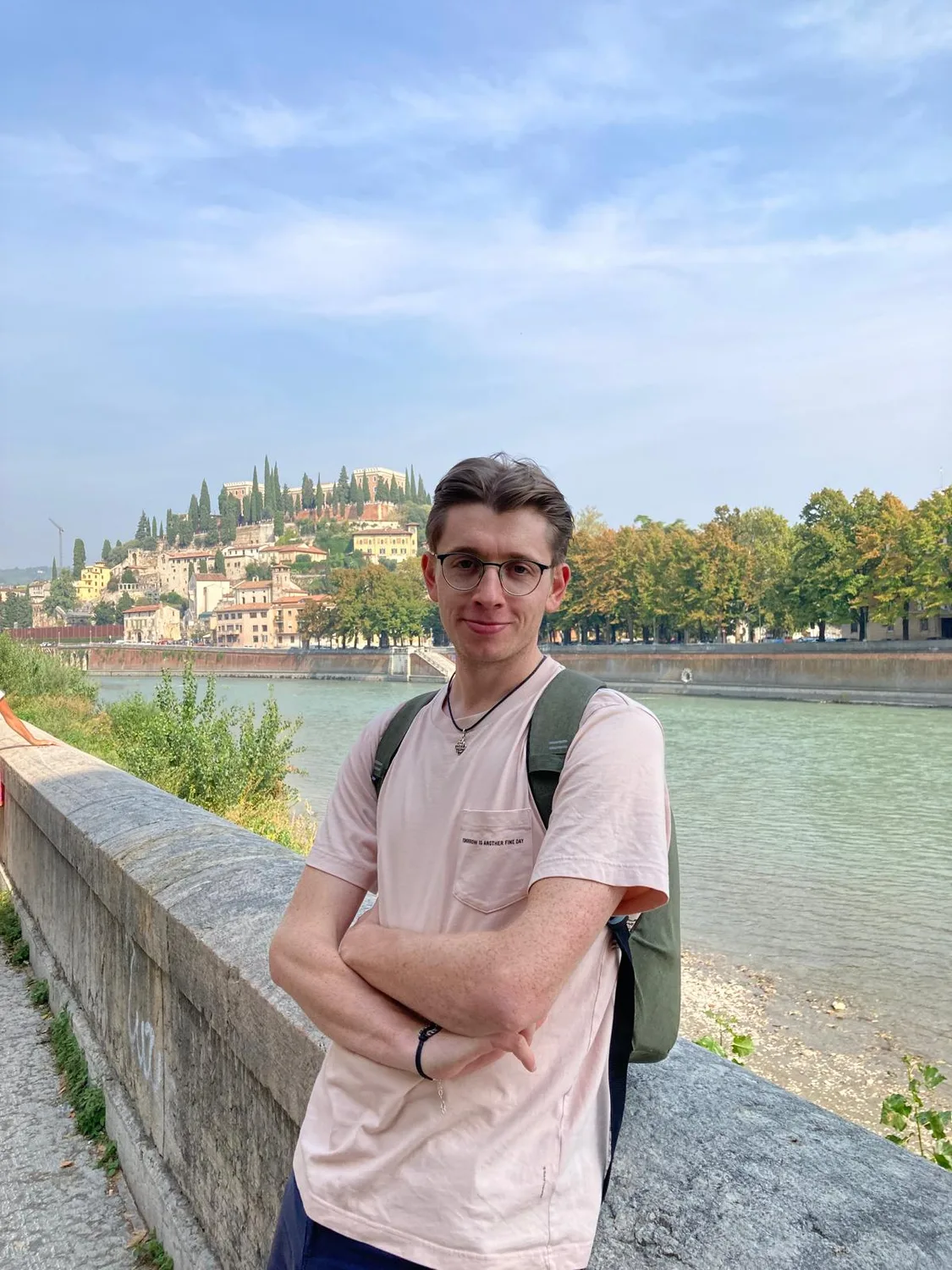 Maximilian Schollemann steht an einer Steinmauer an einem Fluss vor einer italienischen Altstadt mit Kirche und Cypress-Bäume