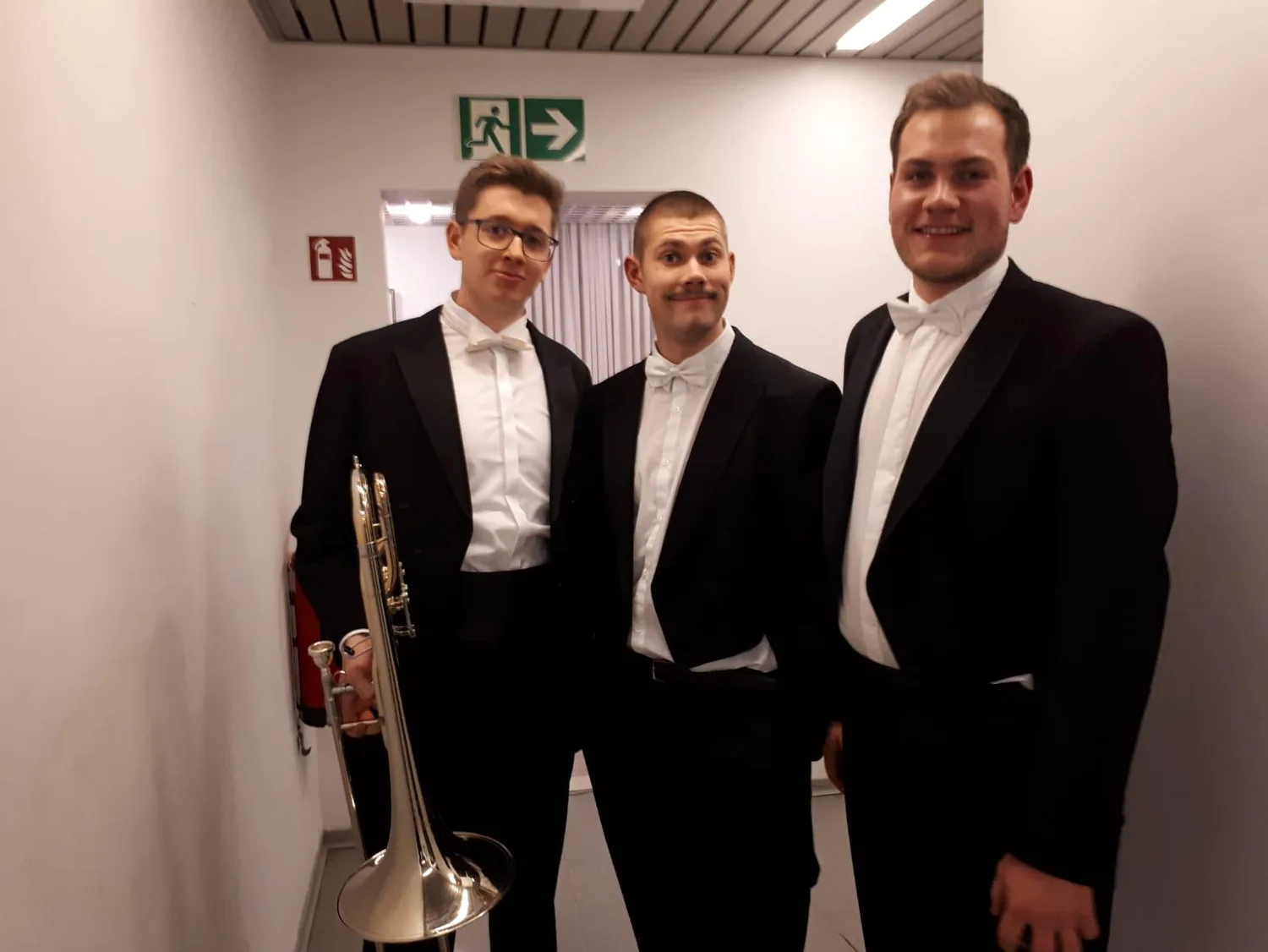 Drei Musiker in schwarzem Anzug und weißem Hemd mit Fliege backstage, linker Person hält Trompete