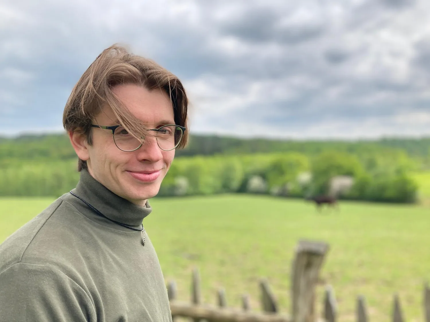 Maximilian Schollemann in grauem Rollkragenpullover und Brille vor grüner Landschaft, Porträt-Foto