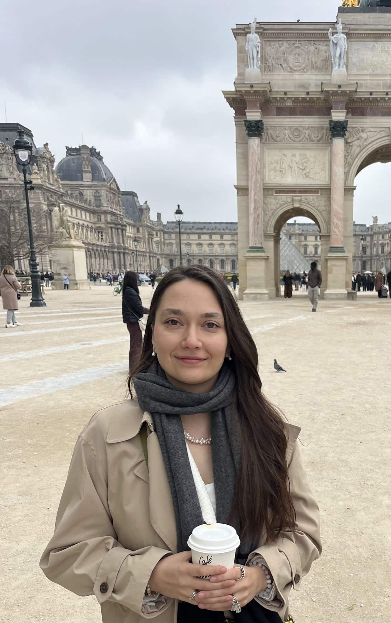 Junge Frau mit dunklen Haaren vor dem Louvre in Paris mit Kaffeebecher, trägt beigen Mantel