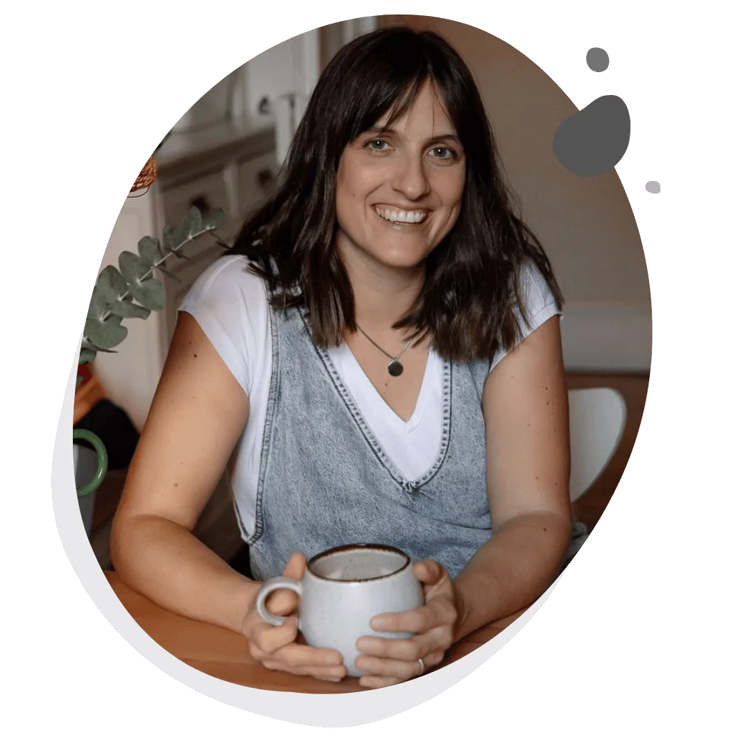Fiona Allan sitzend mit Kaffeetasse, lächelnd, Pflanze im Hintergrund