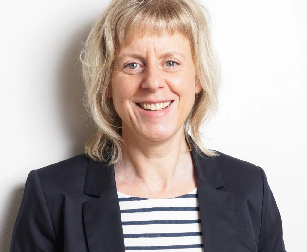 Blonde Frau in schwarzem Blazer mit Streifenshirt lächelnd vor weißem Hintergrund