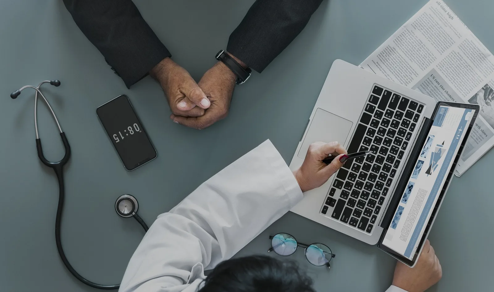 Arzt in weißem Kittel arbeitet am Laptop mit Stethoskop, Handy und medizinischen Unterlagen auf grauem Tisch