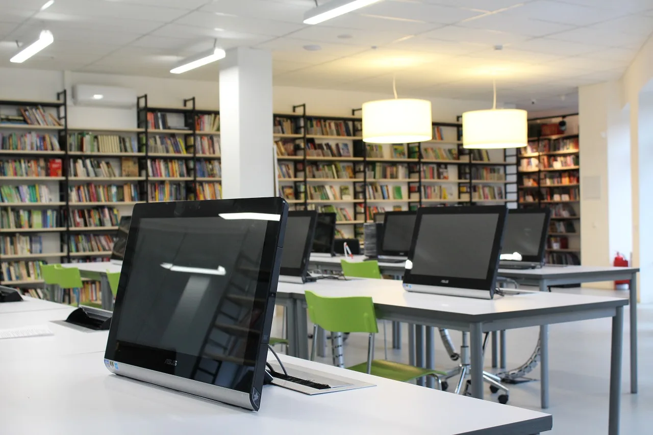 Modernes Schulungslabor oder Computerraum mit mehreren Monitoren, grünen Stühlen und Bücherregalen im Hintergrund
