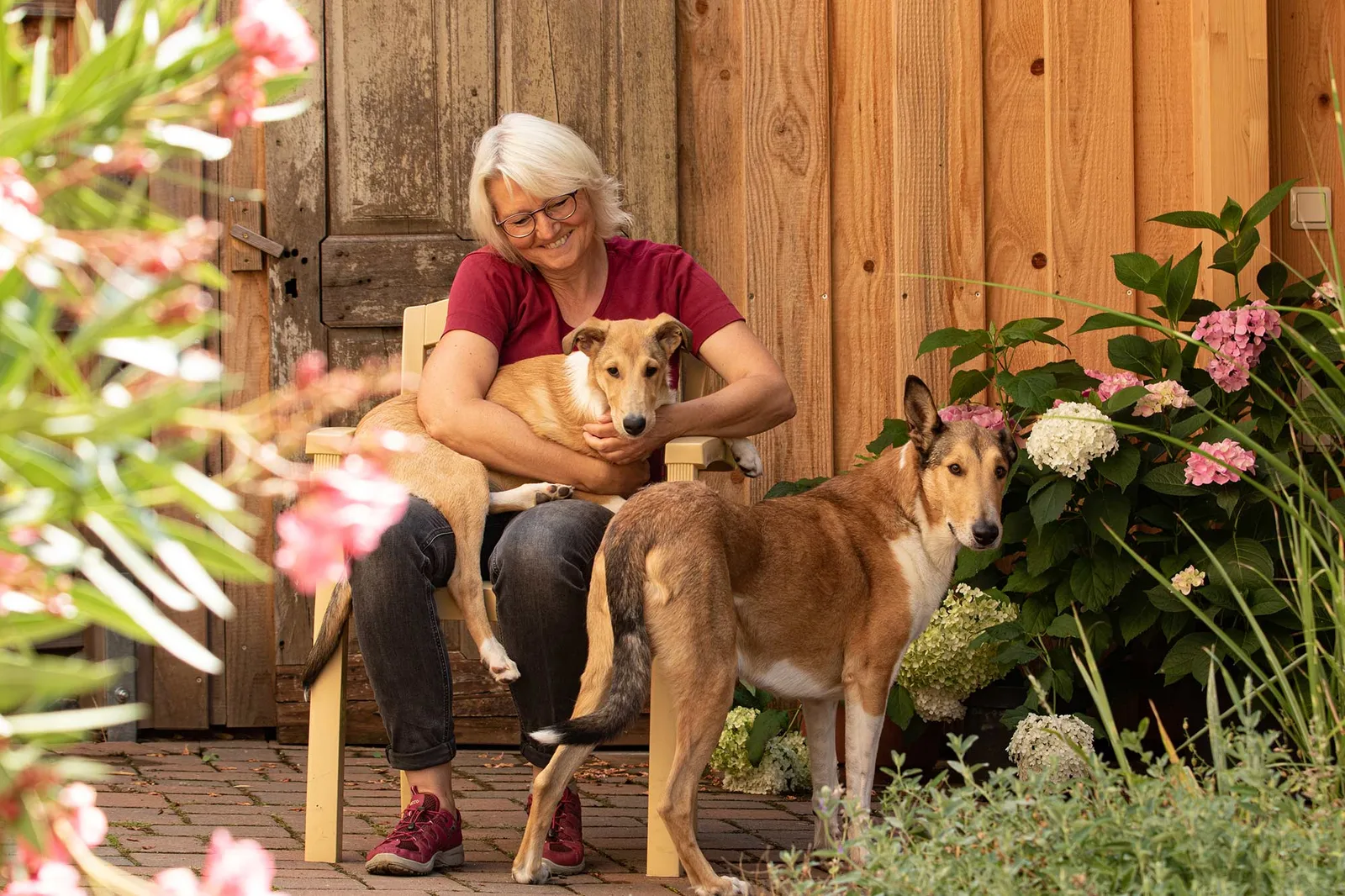 Ältere Frau mit zwei Hunden auf Holzveranda vor Holzwand mit Blüten
