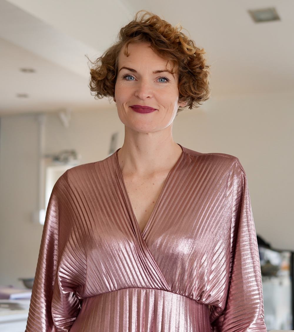 Stefanie Bruns Portrait, Frau mit lockigem rötlich-braunem Haar in rosé glänzendem Oberteil