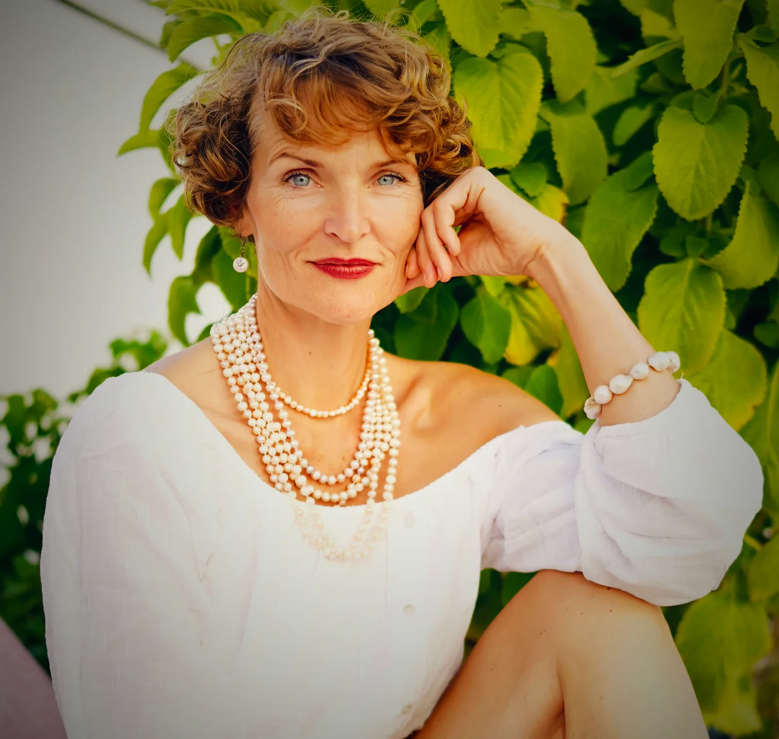 Stefanie Bruns in weißem Oberteil mit mehrreihiger Perlenkette, Hand am Gesicht, grüner Efeu-Hintergrund