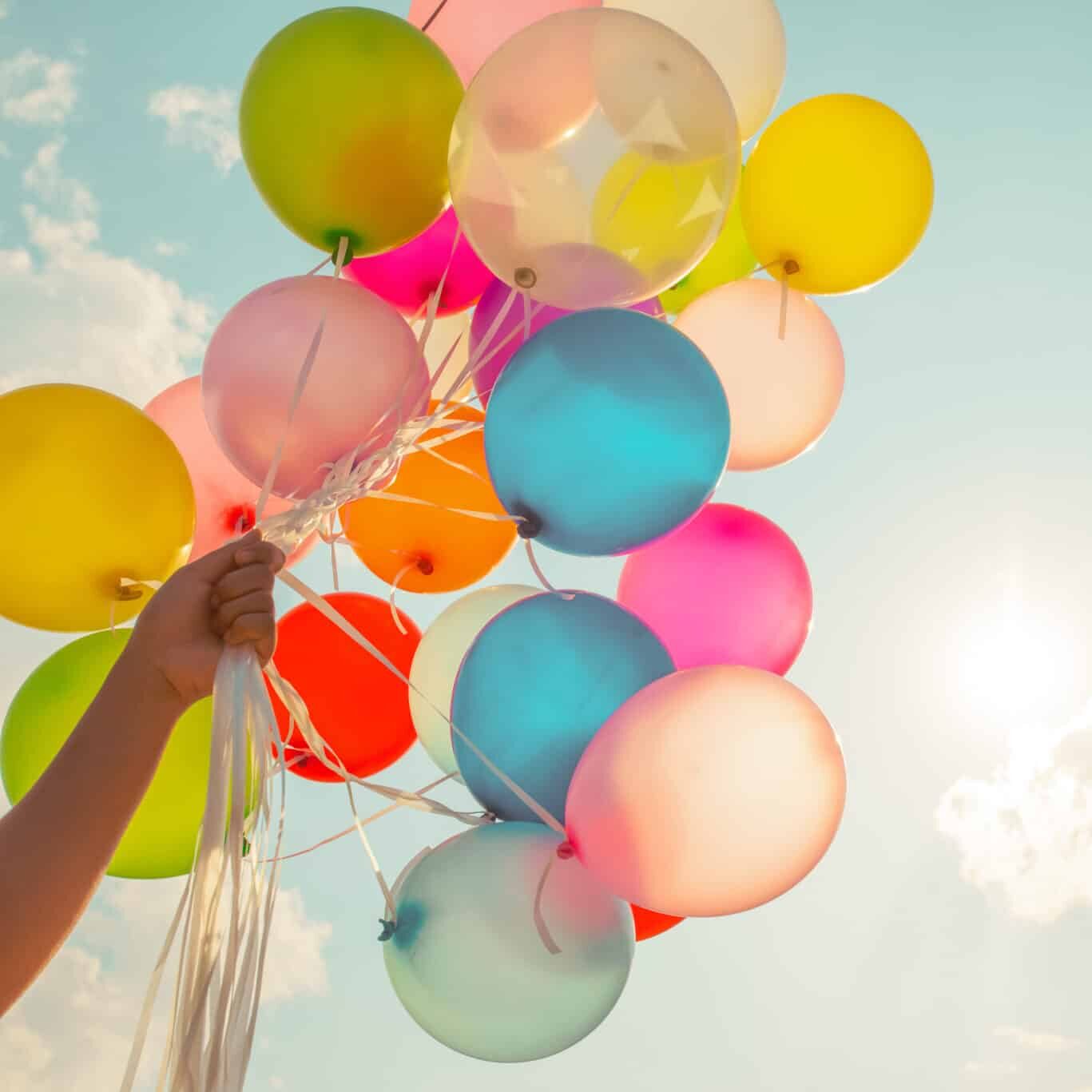 Hand hält bunte Luftballons (gelb, rosa, blau, orange) vor Himmel
