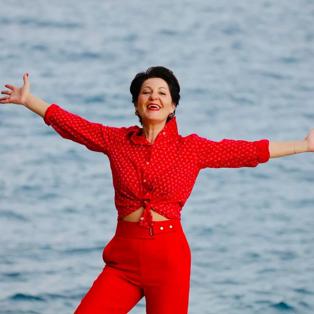 Frau in rotem Outfit mit ausgebreiteten Armen am Meer, lachend, Yoga & Bewusstseinstrainerin