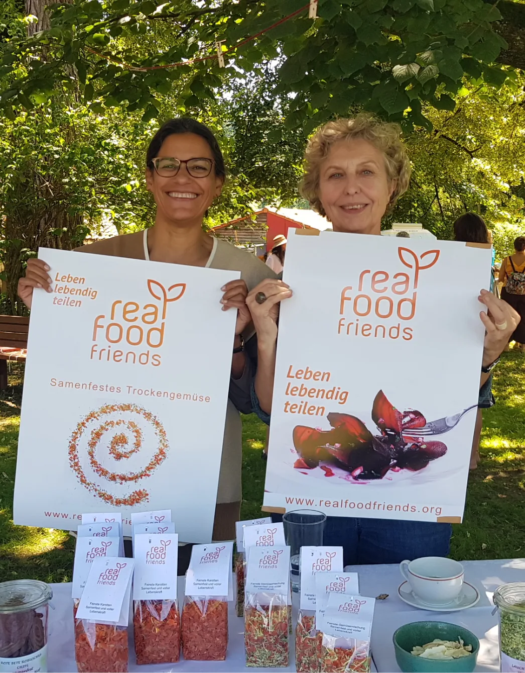 Zwei Frauen mit Real-Food-Friends-Plakaten im Park, Messestand mit Trockengemüse
