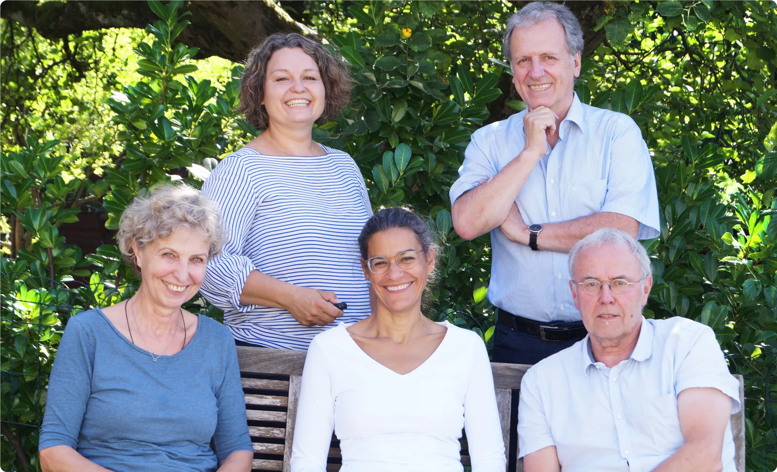 Fünf Team-Mitglieder lächelnd vor grünem Busch-Hintergrund posierend