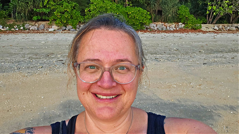 Frau mit Brille und Sonnenbrand am Strand lächelnd – Sicher Alleine Reisen