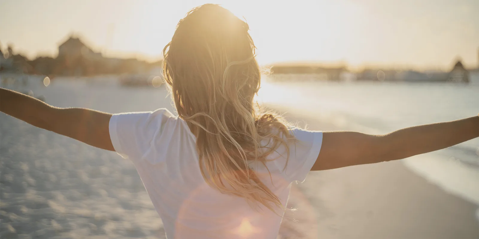 Frau von hinten mit offenen Armen am Strand bei Sonnenaufgang, weißes T-Shirt, lange blonde Haare