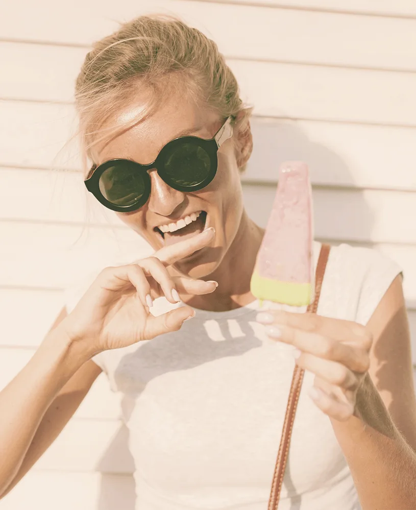 Blonde Frau mit großer Sonnenbrille beim unbeschwerten Genießen von Eiscreme, sommerlich gekleidet