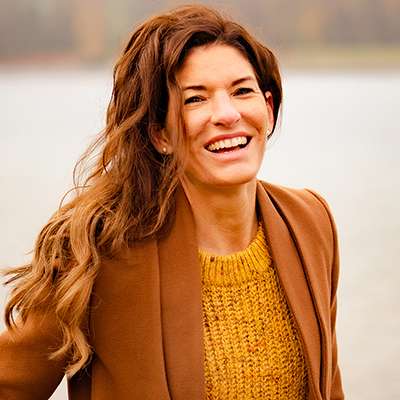 Porträt Rebekka Thiel, rotbraune lange Haare, gelber Pullover, braune Jacke, lachend vor Wasser-Hintergrund