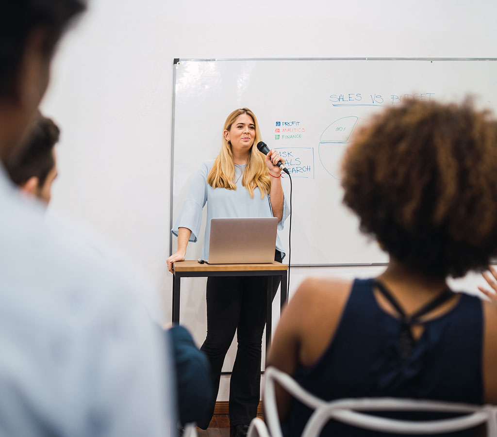 Frau hält Präsentation mit Mikrofon vor Whiteboard mit Business-Diagrammen und Zuhörern