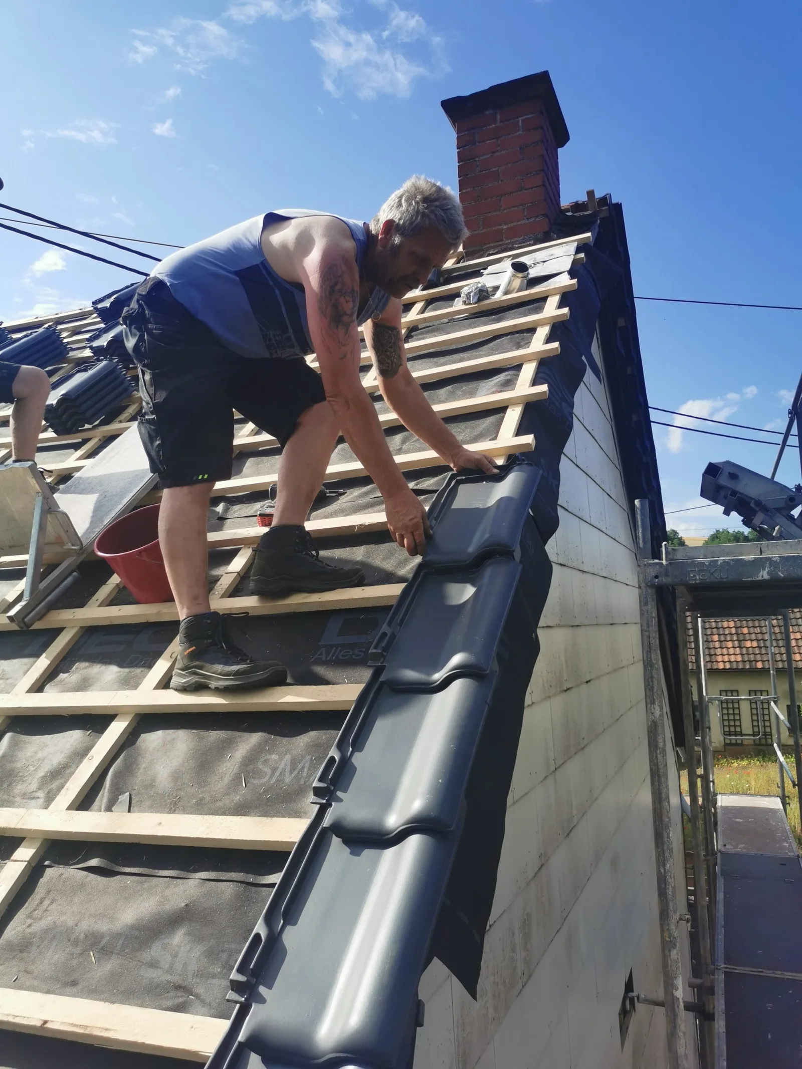 Mann bei der Dachdeckung, kniet auf Dach mit Ziegeln und Werkzeugen, Schritt-für-Schritt Arbeit