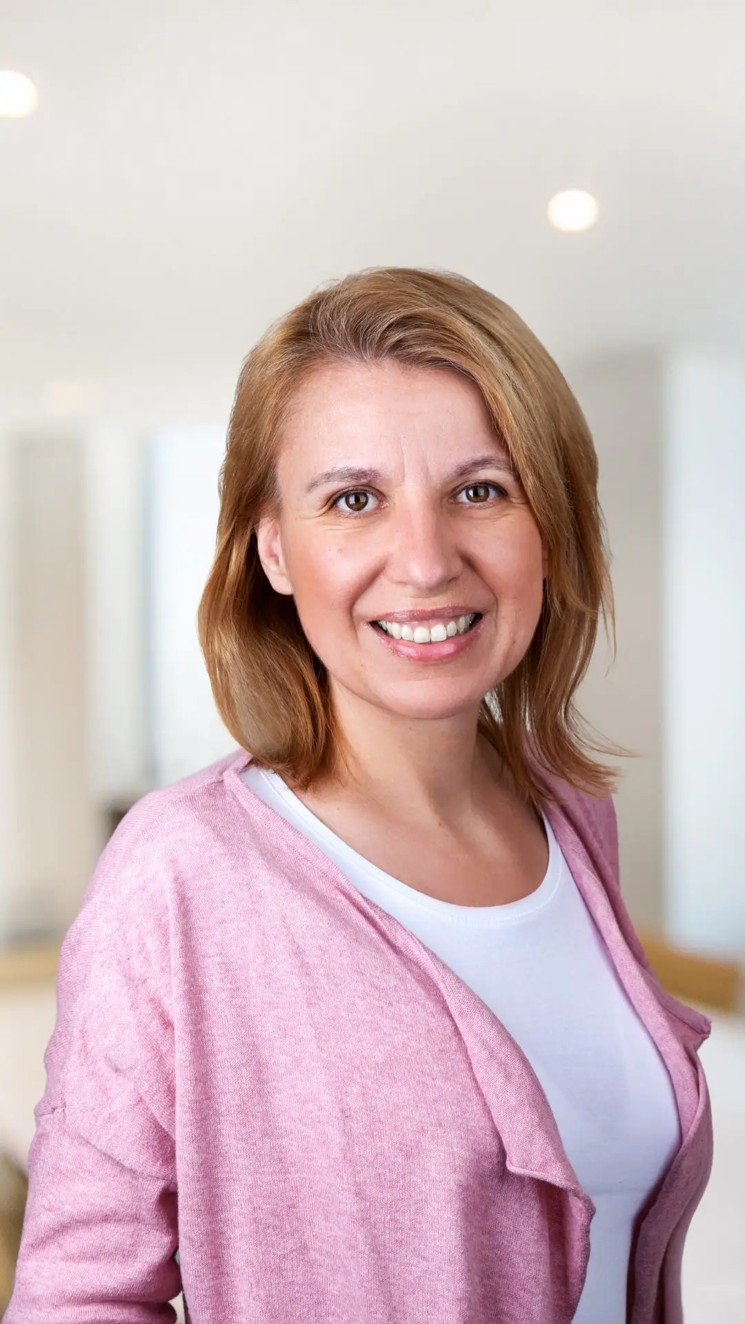 Porträt einer Frau mit schulterlangen rötlich-blonden Haaren, rosa Cardigan über weißem Shirt, lachend
