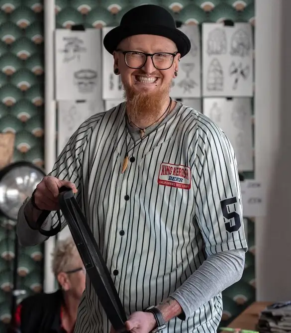 Mann mit rötlichem Bart, schwarzem Hut, Brille, in gestreiftem Baseball-Hemd mit Nummer 5, lächelnd