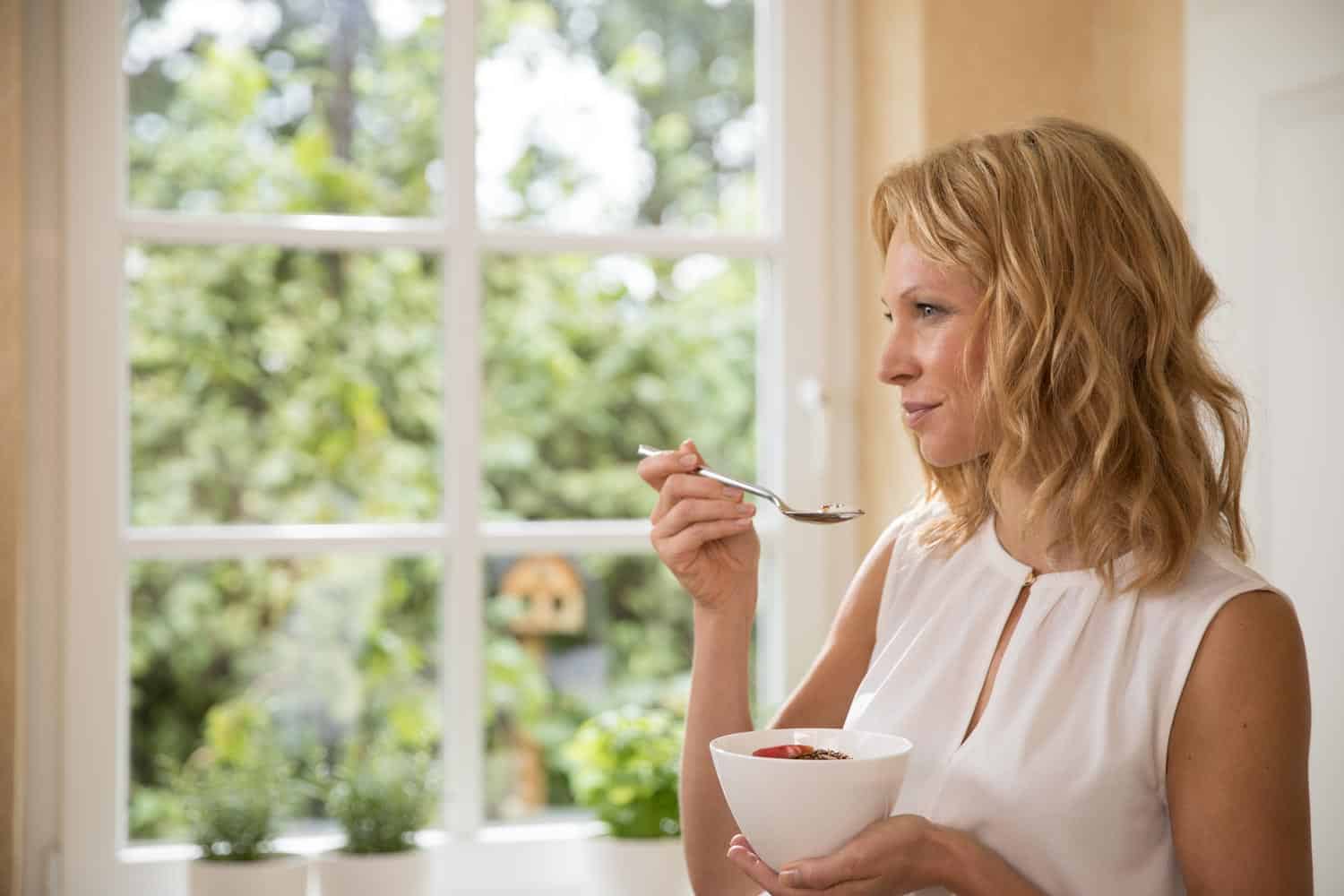 Frau in weißem Oberteil sitzt am Fenster mit Müslischüssel, Ernährung Lifestyle