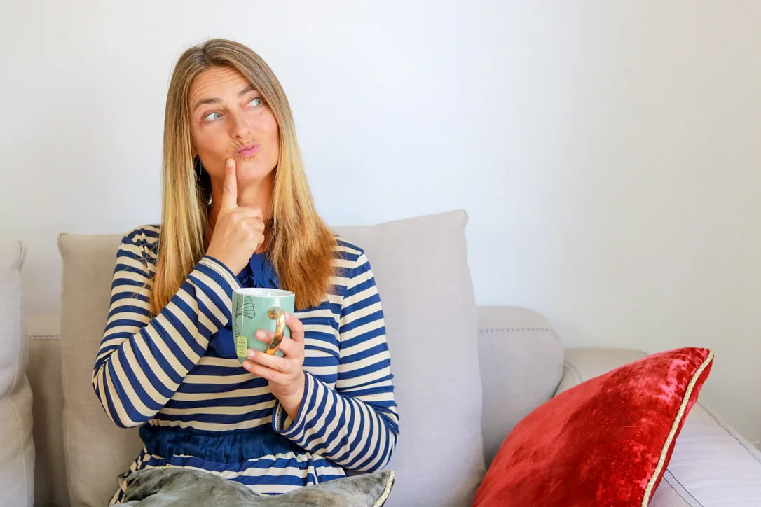 Frau in blau-weiß gestreiftem Blazer sitzt auf Couch mit Kaffeetasse, nachdenklicher Gesichtsausdruck