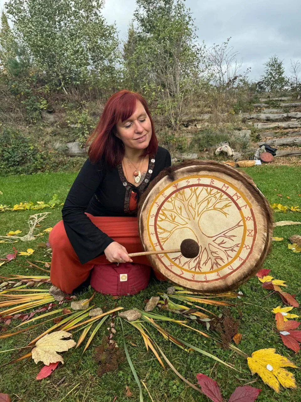 Frau mit roten Haaren hält Holztrommel mit Baumdarstellung und Runensymbolen in Herbstlandschaft