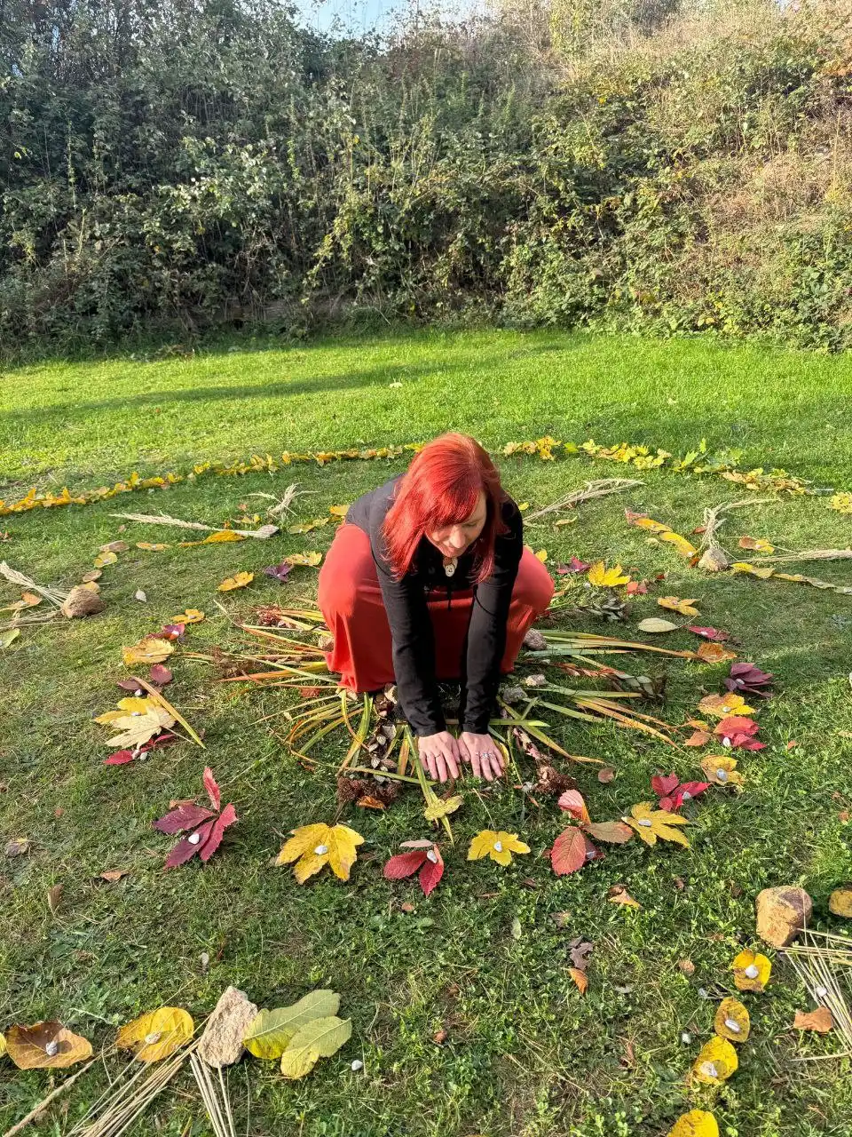 Frau mit roten Haaren sitzt in Herbstkreis aus Blättern auf grüner Wiese