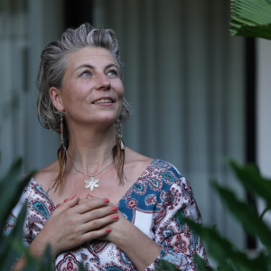 Sacred Woman Rising – Kurspaket heilige Weiblichkeit 41 Porträt von Sandra Hirsch mit ergrautem Haar, Halskette mit Kreuz-Anhänger und gemustertem Oberteil