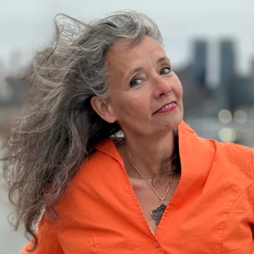 Sacred Woman Rising – Kurspaket heilige Weiblichkeit 28 Christiane Trautwein-Lykke mit grauem Haar in orange Jacke vor Stadtkulisse