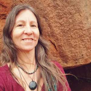 Sacred Woman Rising – Kurspaket heilige Weiblichkeit 34 Porträt von Sibylle Moana mit braunem Haar, Halskette und pink-rotem Oberteil vor braunem Hintergrund