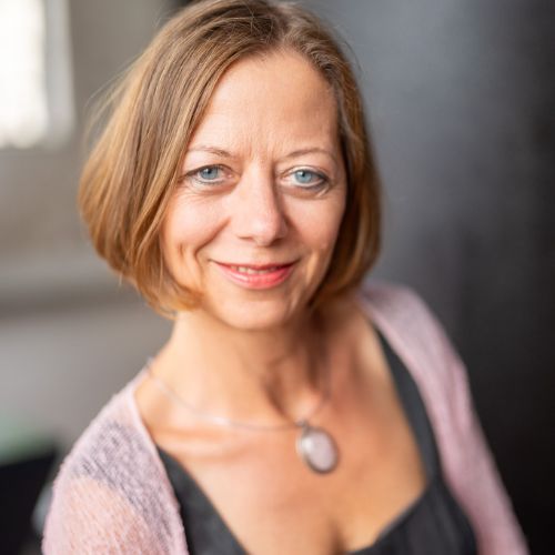 Sacred Woman Rising – Kurspaket heilige Weiblichkeit 35 Porträt von Silke Virajá Göbel mit braunem Haar, grauem Oberteil und grauem Anhänger-Schmuck