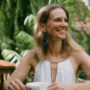 Sacred Woman Rising – Kurspaket heilige Weiblichkeit 21 Porträt von Julia Mandel mit langen blonden Haaren und Ohrhängern vor grünem Laub lächelnd