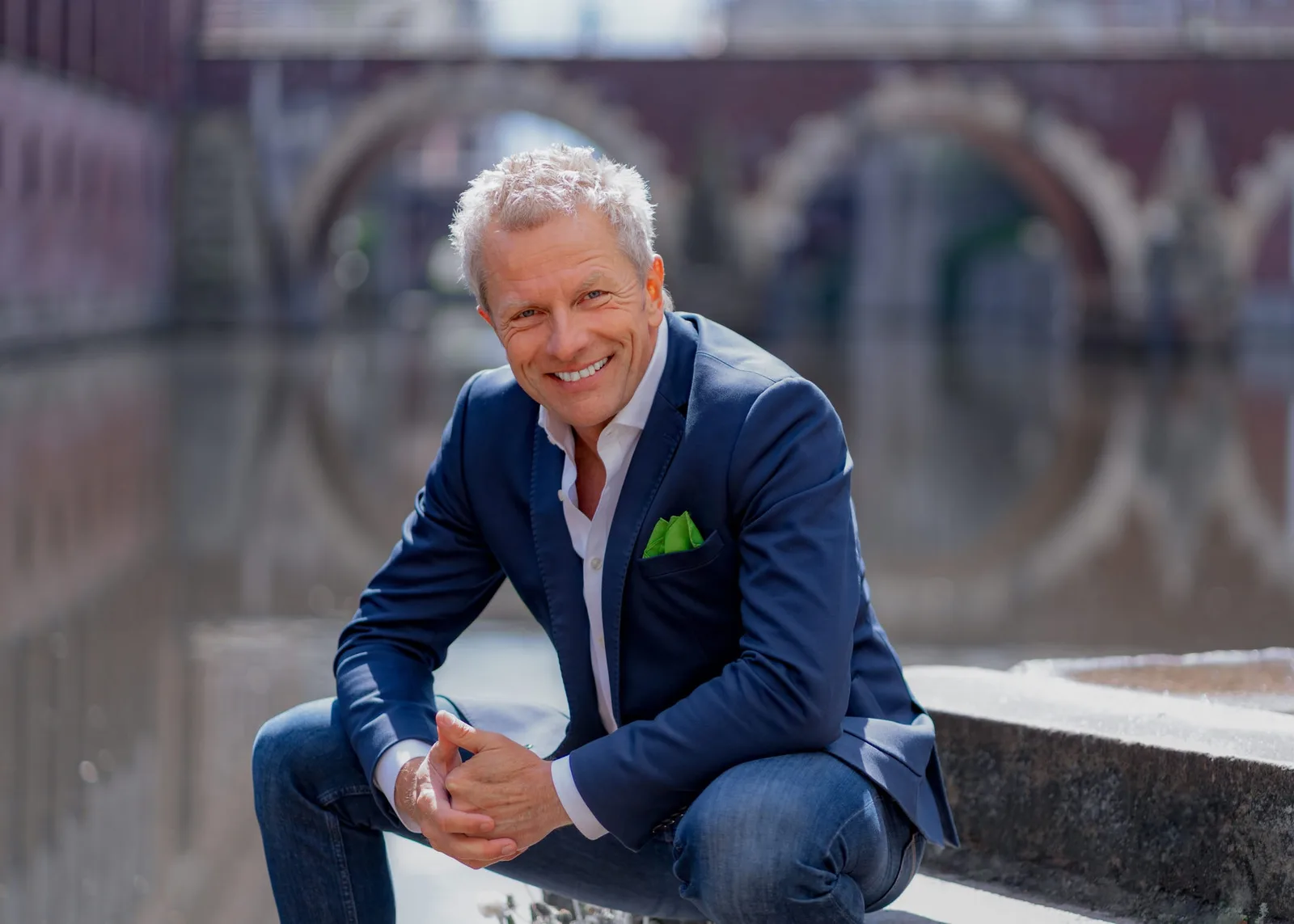 Andreas Enrico Brell, älter Mann mit weißem Haar in dunkelbauem Blazer und blauem Hemd vor unscharfem Hintergrund