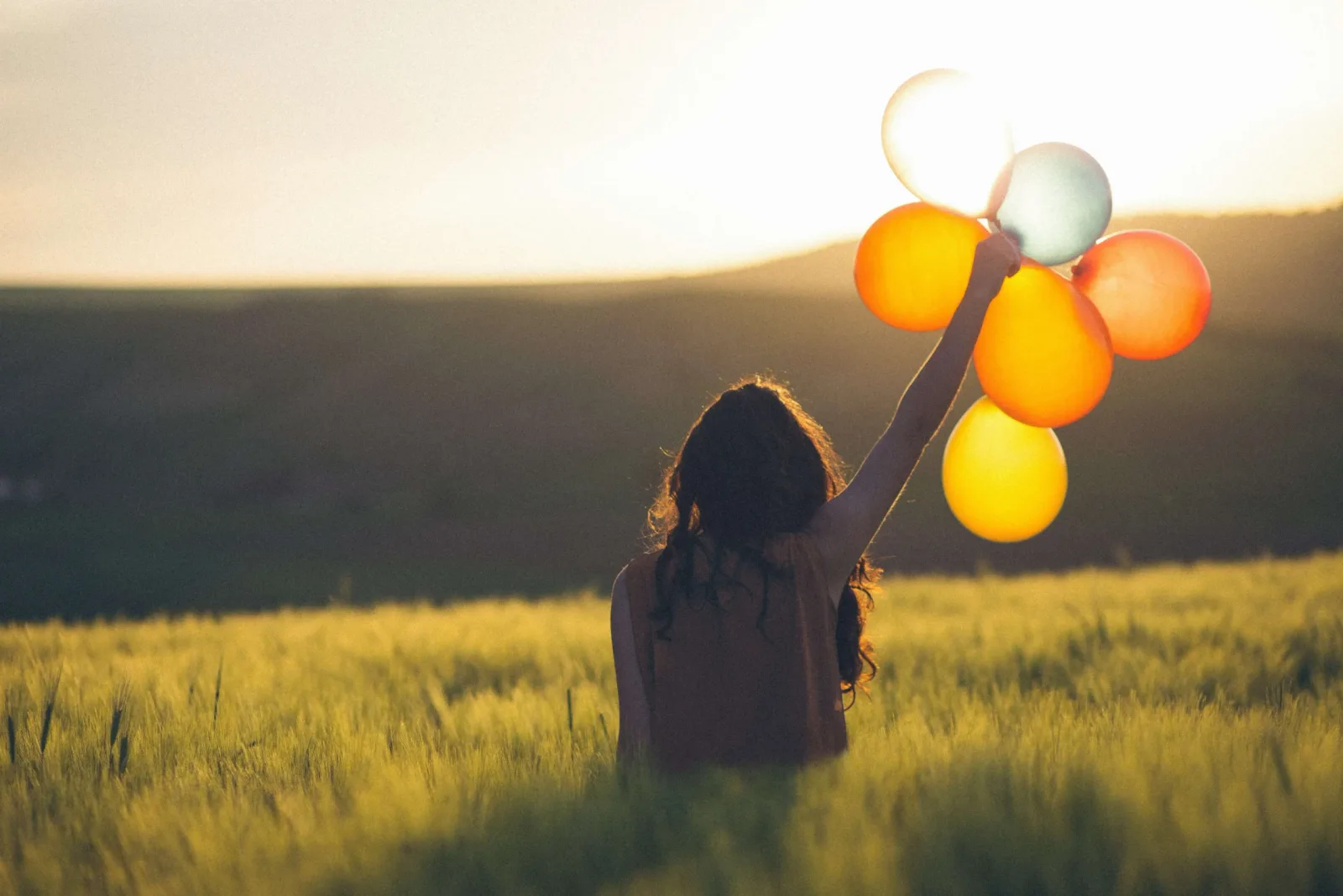 Frau von hinten in Getreidefeld bei Sonnenaufgang, hält bunte Luftballons in die Luft