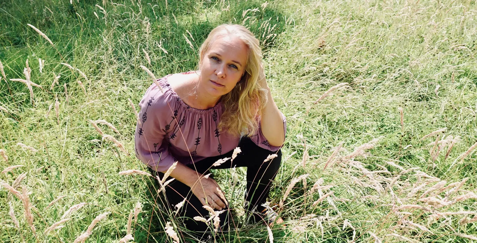 Blonde Frau in rosafarbener Bluse kniet in trockener Wiese, nachdenklicher Blick zur Seite