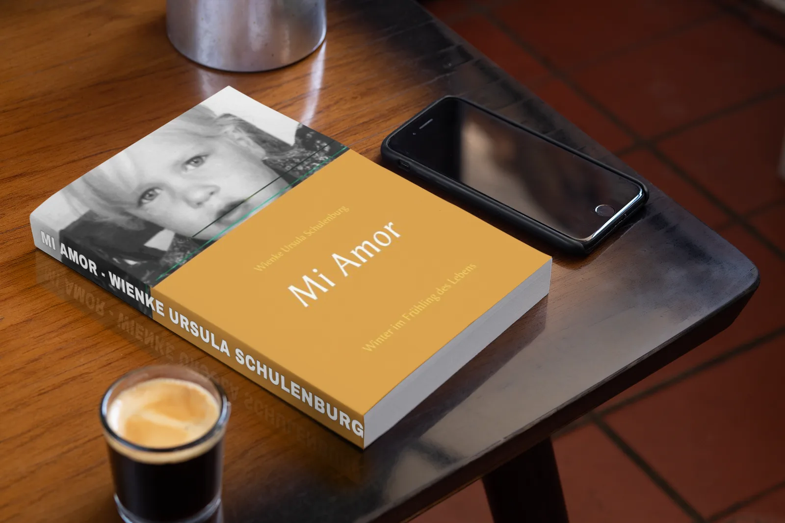 Buchcover Mi Amor in Gelb neben Schwarzweiß-Porträt, auf Holztisch mit Smartphone und Kaffee