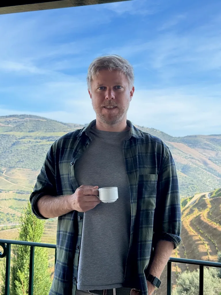 Tim Hamer in kariertem Hemd mit Kaffeetasse vor Weinbergslandschaft