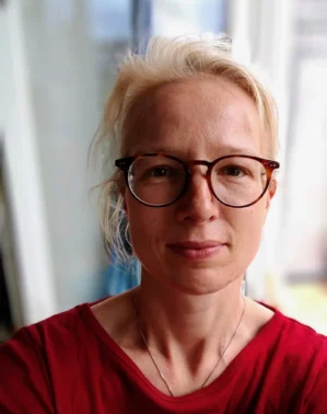Porträt von Pia Helfferich mit Brille, rötlichblondem Haar im Dutt, rotes Shirt