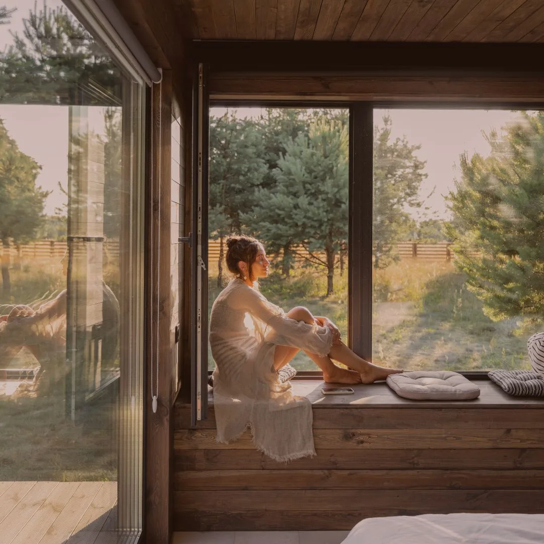 Frau sitzt am Fenster mit Blick auf Wald und Wiese, Morgen- oder Abendlicht