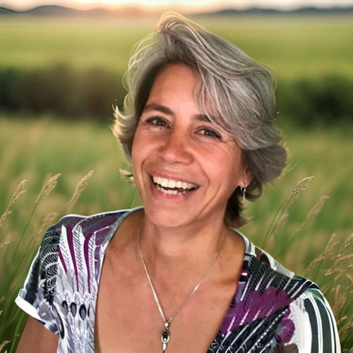 Silke Busse mit grauem Haar in lila-weißem Muster-Oberteil vor Getreidefeld, lächelnd