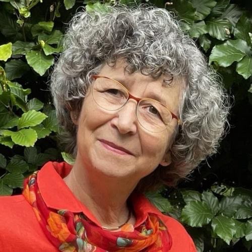 Porträt Karin Reschke, ältere Frau mit grauem lockigem Haar, Brille, rotem Oberteil, grüner Blatterhintergrund