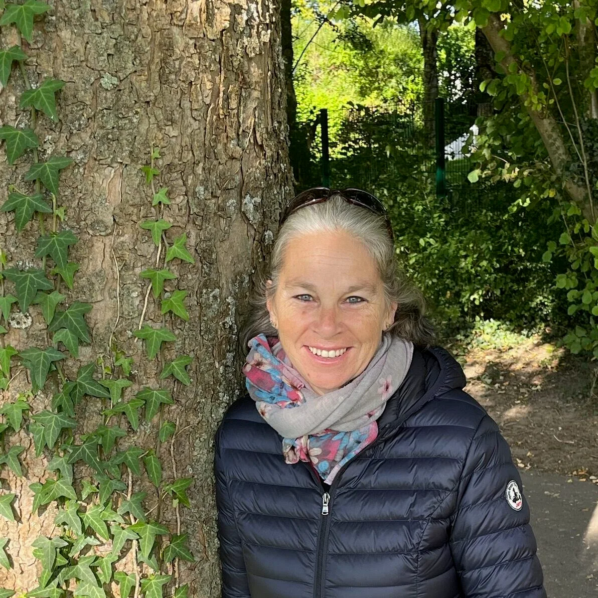 Doris Forster vor Baumstamm im Wald mit Schal und schwarzer Jacke, lächelnd