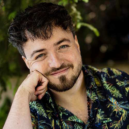 Porträt Enrico Fricke, Mann mit schwarzem Haar, beigem Blumenshirt, Hand am Kinn, grüner Hintergrund