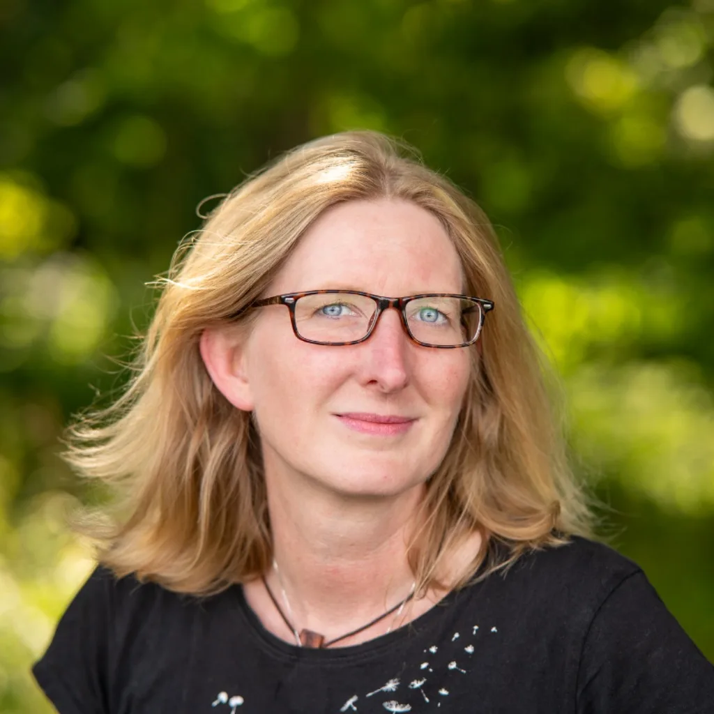 Portraitfoto einer Frau mit rötlich-blondem Haar, Brille und schwarzem Oberteil vor grünem Hintergrund