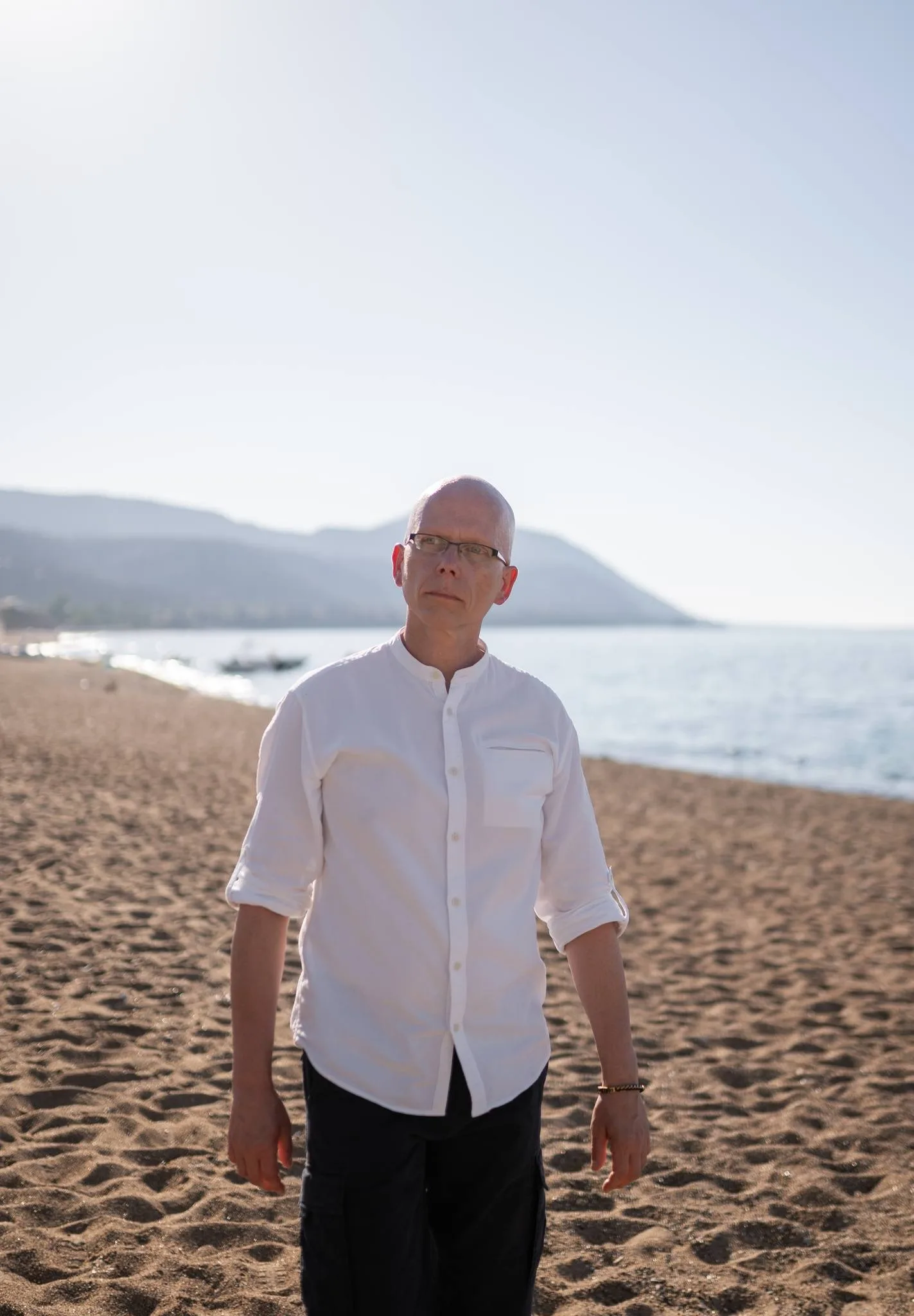 Mann mit Glatze und Brille in weißem Hemd an einem Strand mit Bergen im Hintergrund
