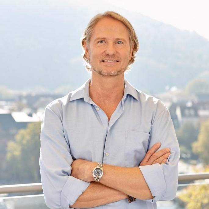 Porträt von Steffen Lohrer, Mann mit blondem Haar in hellblauem Hemd, verschränkte Arme, Stadtlandschaft im Hintergrund