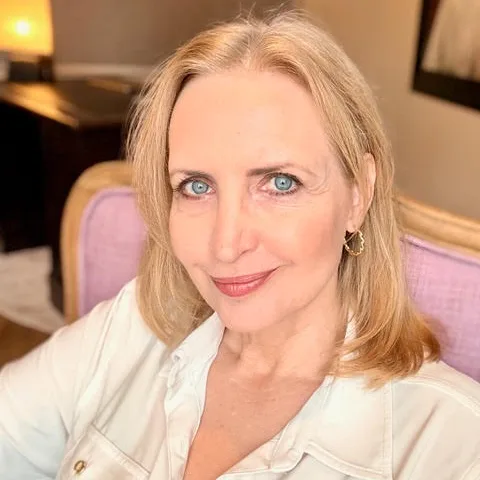 Porträt Martina, Frau mit hellbraunen mittellangen Haaren, weißem Top, lila Stuhl, warmer Innenraum-Hintergrund