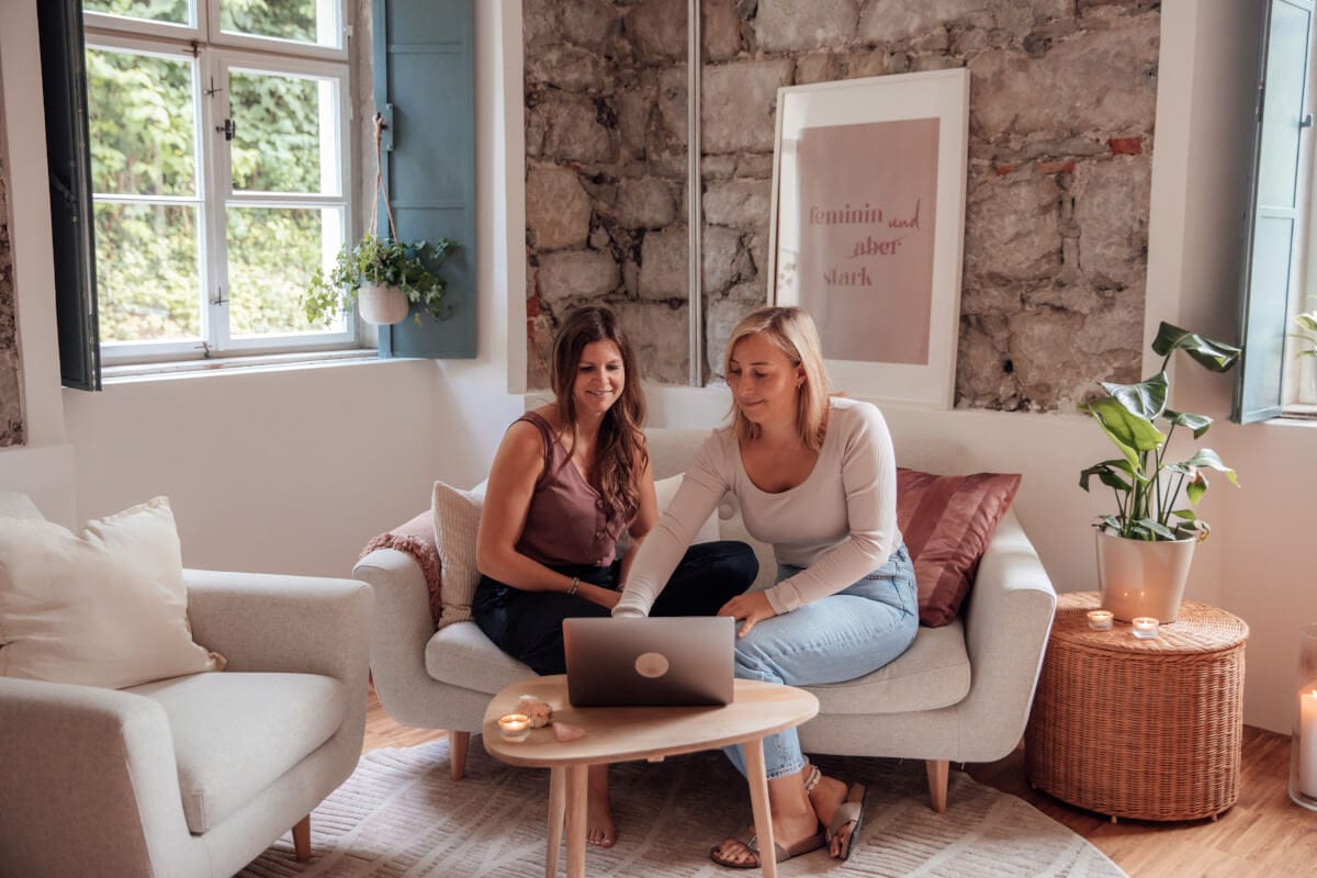 Jana und Lisa sitzen auf einem Sofa und besprechen gemeinsam am Laptop die Kursinhalte