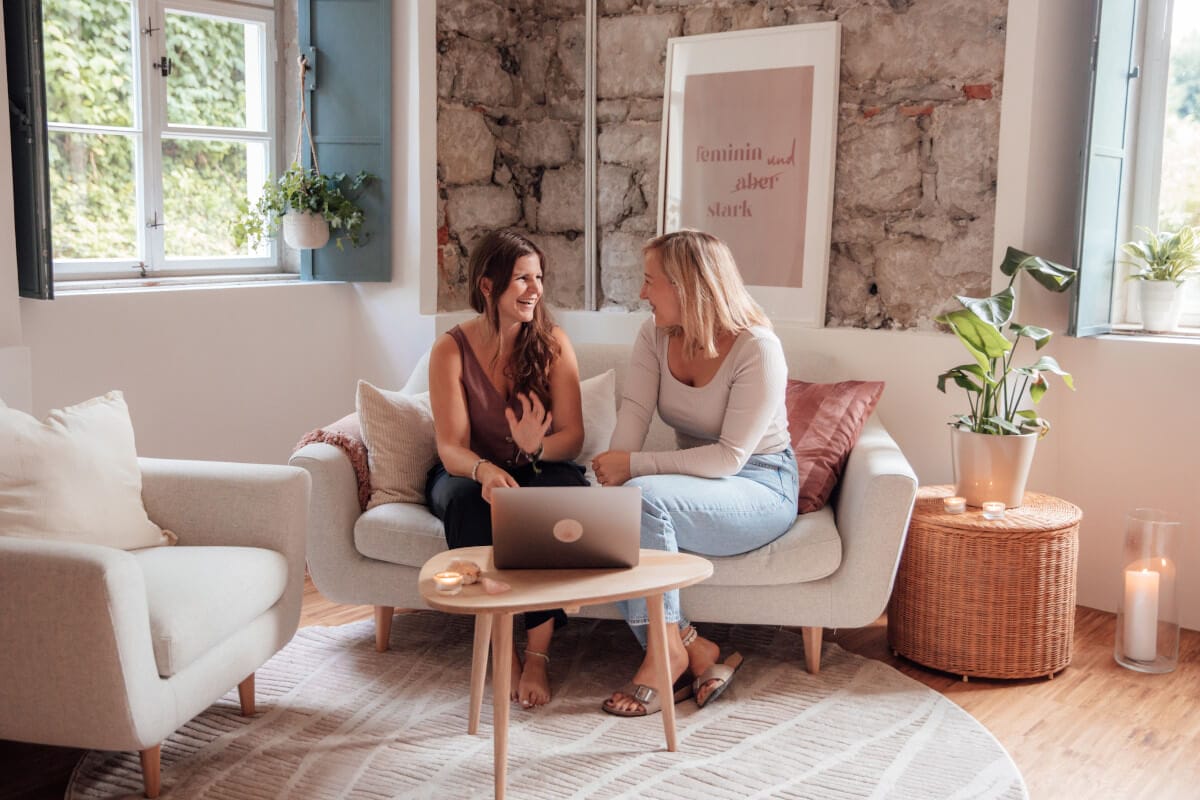 Jana und Lisa sitzen auf einem Sofa und unterhalten sich über die exklusive Community für Business-Frauen