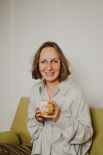 Porträt von Linda Klein mit neutralem Hintergrund, lächelnd mit Becher